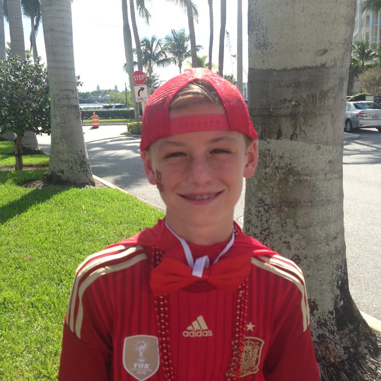 Image of young boy wearing all red. Smiling towards the camera. The scenery is of a tropical residential area and palm trees can be seen in the background. The boy is wearing a hat, face paint, a jacket, a bowtie, and beaded necklaces all in red. 
