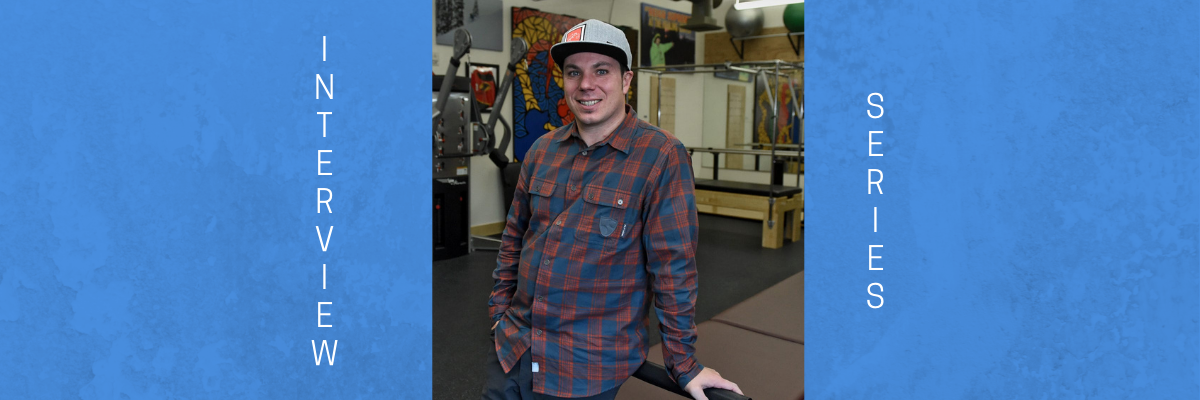 Image of Roy Tuscany. He is wearing a blue and red plaid shirt and jeans. His right hand is in his pocket. His left hand is on a table, which he is leaning against. He is Caucasian and is wearing a baseball hat and smiling at the camera. Roy has dark brown hair, which is protruding out of his hat. The background is of a physical therapy studio.