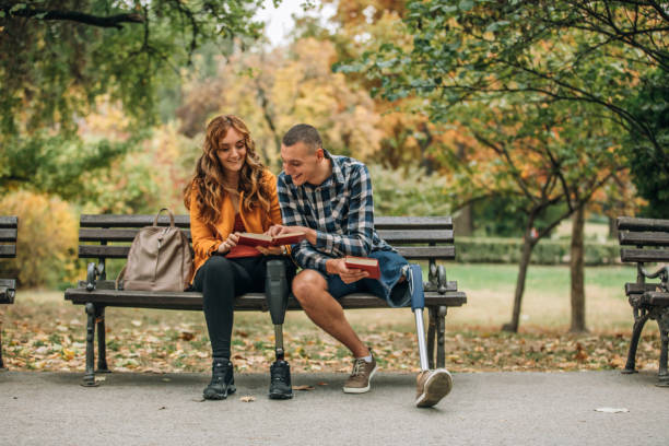 Two people sit on a bench. Girl and guy in early 30s. They look romantic on a bench with a park scene behind them. They both are missing legs and show prosthetics.