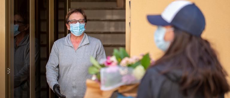 Image Description: A man, wearing a mask and gloves, receives a flower delivery from a woman also wearing a mask and gloves. 