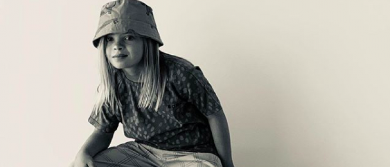 Image Description: Sofia Sanchez poses for a modeling shoot. She is seen in black and white. A bucket hat covers her long blond hair. She leans her right hand on her knee and offers a serious, cool look to the camera. 