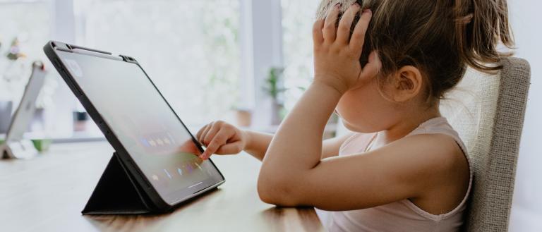 Child (girl) sitting at a table with a flat screen lap top. Her hand is on her head signaling stress.