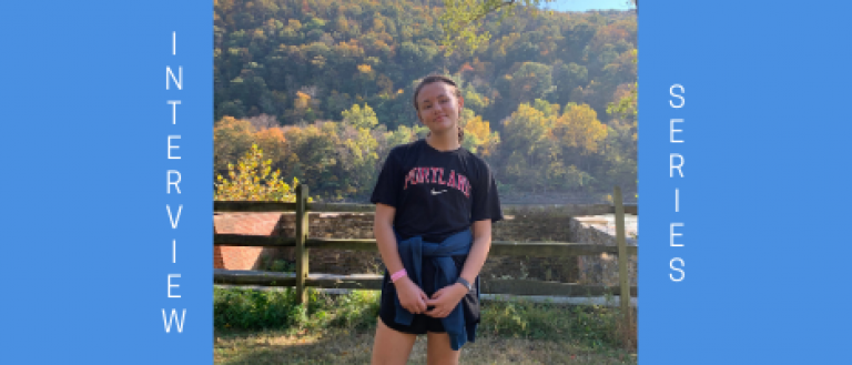 Image of teenage girl near a wooden fence outside. She is smiling, wearing shorts and a t-shirt. There is a fall foliage background.