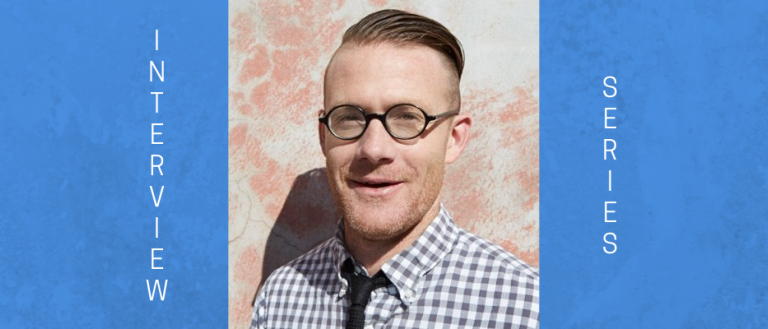 Image of man smiling at the camera. He is wearing a white and blue small checkered shirt and a blue tie. He is wearing glasses and his hair is parted to the side.