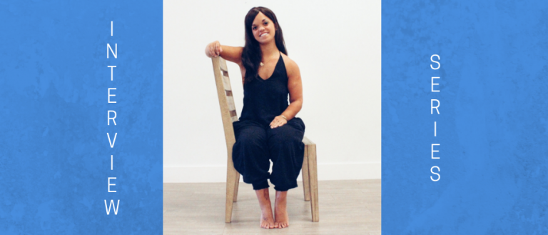 Image of woman sitting on a chair sideways, smiling towards the camera. She has long brown hair and is wearing a black romper.