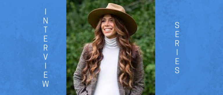 Image of a woman wearing a white turtleneck, a brown blazer, and a wide brim brown hat, She has long hair and is smiling towards the left of focus. There are pine trees behind her. 