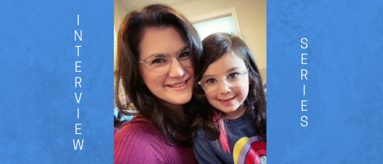 Image of mother and 4 year old daughter. Daughter is sitting on her mother's lap. They are both wearing glasses and smiling at the camera. 