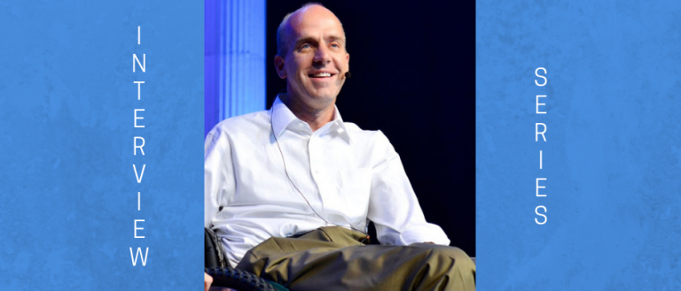 Image of Chad Hymas. He is sitting in a wheel chair in a white dress shirt and brown pants, who is bald. The background is a lighted stage, as he is presenting as a motivational speaker. Chad is wearing an over the ear microphone.