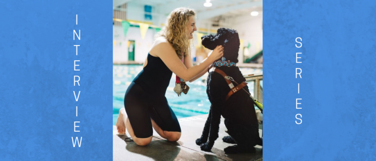 Image of Olympic swimmer McClain Hermes. She is wearing the medals that she has won. She is bending down petting her seeing eye dog, which is black and medium sized. They are in front of an indoor pool. McClain is looking at her dog and smiling.