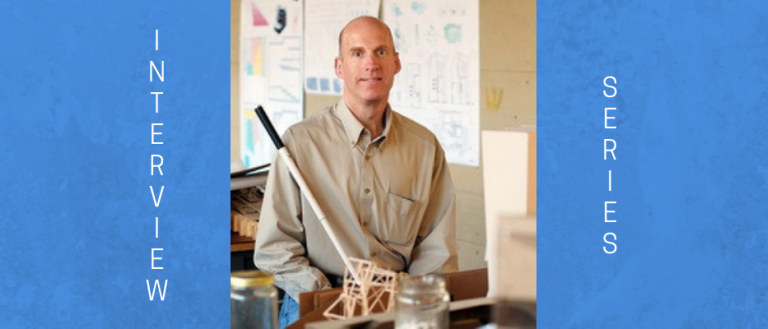 Image of Chris Downey, an architect who is blind. He is bald and wearing a tan colored shirt and jeans. He is holding his walking cane and smiling at the camera. Behind Chris is an architect workshop, filled with prints/plans, building objects/tools, and 3-D models of buildings. 