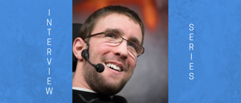 Headshot image of Mike Berkson, comedian with cerebral palsy. His is looking up and to the right. He is wearing glasses, a black shirt with a white undershirt. He has a clean-cut look with his hair, beard, and moustache well kempt. He is wearing a headset/microphone as he is speaking to an audience during one of his motivational speaking engagement.    