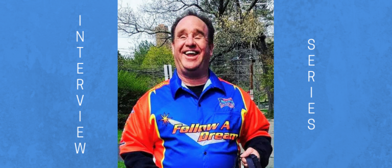 Image of Jay Blake. He is wearing a sports racing jacket that reads "Follow A Dream," which is blue, orange and white. He has a big smile and short brown hair. Jay is also blind. The background consists of trees with park scenery.