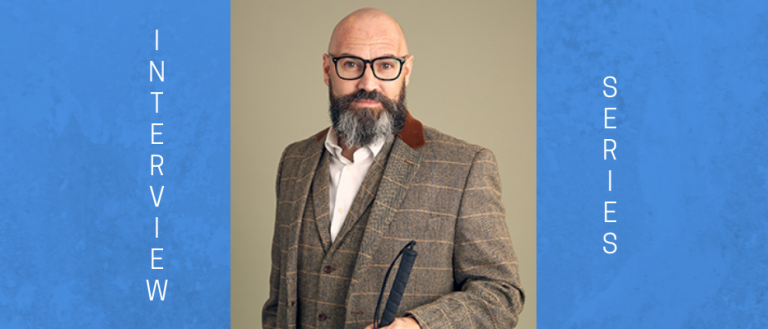 Image of Dave Steele. He is in front of a tan backdrop. He is a bald man. He is wearing thick black glasses and has a gray and black beard/moustache combo. He is wearing a brown tweed print suit with vest and a white collard shirt that is unbuttoned at the top. He is holding his walking stick and facing the camera.