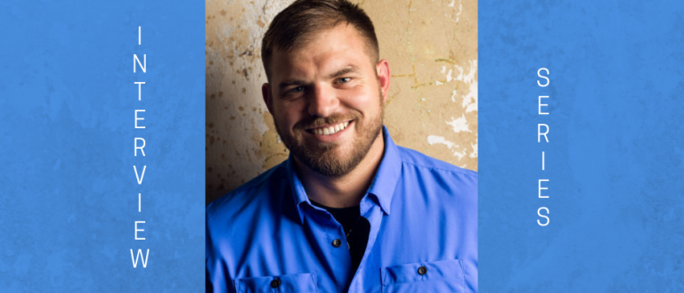 Image of Travis Mills. The image is in portrait mode with the background being abstract in tan and white. Travis is smiling at the camera; he is wearing a blue jean button down shirt with the top button open, which exposes a black t-shirt. He is of a stockier build and has short military cut brown hair and a brown beard/moustache combo.  