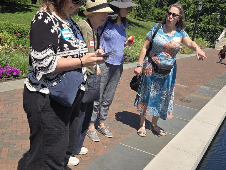A group of four people observes a water feature, with one person gesturing while explaining something. They are outdoors in a park on a sunny day.