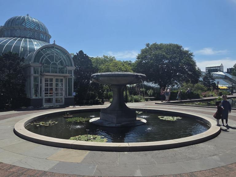 A circular fountain surrounded by stone pathways and greenery, with a glass conservatory in the background under a clear blue sky.