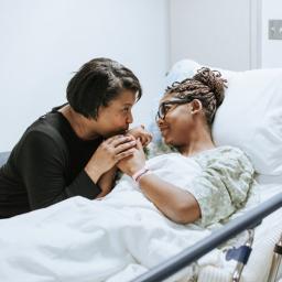 Two women hold hands and look at one another. One is in a hospital bed, wearing a hospital gown. 