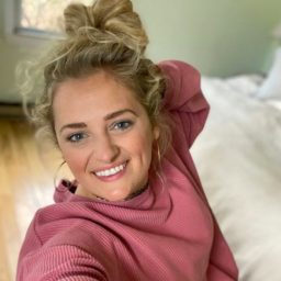 Ali Stroker is pictured with one hand behind her head. She is wearing a pink sweater and flashing a big smile for the camera. 