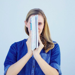 Image Description: Kathryn faces the camera, wearing a blue shirt and a smile. In front of her face, however, she holds her book (from the side) so that it obscures half her face. 
