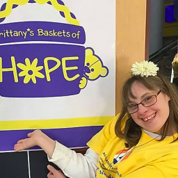 Image Description: Brittany Schiavone poses with the Brittany's Baskets of Hope logo. She wears a yellow t-shirt and has a white flower in her hair. 