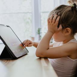 Child (girl) sitting at a table with a flat screen lap top. Her hand is on her head signaling stress.
