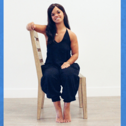 Image of woman sitting on a chair sideways, smiling towards the camera. She has long brown hair and is wearing a black romper.
