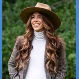 Image of a woman wearing a white turtleneck, a brown blazer, and a wide brim brown hat, She has long hair and is smiling towards the left of focus. There are pine trees behind her. 