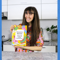 Image of author Amanda Orlando holding her cookbook Everyone's Welcome in her kitchen. She is wearing a patterned dress and her brown hair down. She also has bangs that are slightly parted. She is smiling at the camera.
