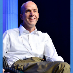 Image of Chad Hymas. He is sitting in a wheel chair in a white dress shirt and brown pants, who is bald. The background is a lighted stage, as he is presenting as a motivational speaker. Chad is wearing an over the ear microphone.