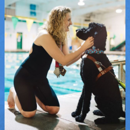 Image of Olympic swimmer McClain Hermes. She is wearing the medals that she has won. She is bending down petting her seeing eye dog, which is black and medium sized. They are in front of an indoor pool. McClain is looking at her dog and smiling.