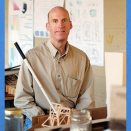 Image of Chris Downey, an architect who is blind. He is bald and wearing a tan colored shirt and jeans. He is holding his walking cane and smiling at the camera. Behind Chris is an architect workshop, filled with prints/plans, building objects/tools, and 3-D models of buildings. 