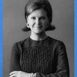 Black and white image of Laura Slatkin, CEO of NEST Fragrances and Founder of NEXT for AUTISM. Laura is wearing a long sleeved, black blouse with small ruffles in the center that are vertical. She is wear in two bracelets, a ring on her left hand, and earrings in a hoop shape. Her hair is in bouffant  style. She is facing forward in the photo with her arms crossed and smiling.