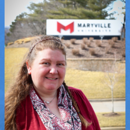 Image of Katie Selby. She is positioned left of the photo wearing a red sweater and a colorful scarf. Her brown hair is pushed back using a headband. She also has a braid on the left side of her head She is wearing a silver necklace and is smiling in the photo. She is standing In front of a Maryville College sign, which is in the distance behind her on a grassy hill. 