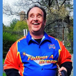 Image of Jay Blake. He is wearing a sports racing jacket that reads "Follow A Dream," which is blue, orange and white. He has a big smile and short brown hair. Jay is also blind. The background consists of trees with park scenery.