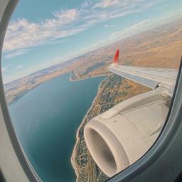 Image of inside airplane window showing a birds eye view of undisclosed land and water below; it is angled and the airplane wing and engine is also featured. The sky in the photo is in daytime mode and there are several cirrus clouds floating.  