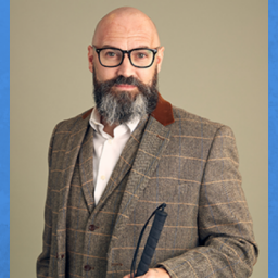 Image of Dave Steele. He is in front of a tan backdrop. He is a bald man. He is wearing thick black glasses and has a gray and black beard/moustache combo. He is wearing a brown tweed print suit with vest and a white collard shirt that is unbuttoned at the top. He is holding his walking stick and facing the camera.