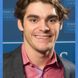 Image of RJ Mitte. It is a headshot of him in front of a blue background. He is smiling at the camera. RJ is wearing a light red button up shirt and a gray sports coat. the top button of the shirt is undone. RJ has thick brown eyebrows and a brown short hair that is a bit unkempt looking. He is 29, but visually looks in his early 20s/Late teens.  