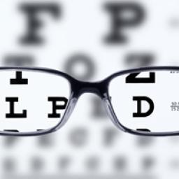 Image of an eye test chart which is blurry. There are black reading glasses superimposed on the image and through those lenses the letters L, D, P, and the number 4 can be seen.