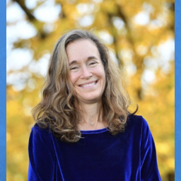 Image of Michelle Bernstein. She is a woman in her 50s, Caucasian, and long dirty blonde gray hair. In the photo she is wearing a boatneck top in cobalt blue velvet. The photo is from the chest up. The background is blurred with yellow leaves on a tree. 