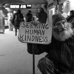 Homeless man in subway station holding a sign that says, "Seeking Human Kindness." The photo is in black & white.
