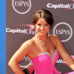 Image of Victoria Arlen at the ESPYS red carpet event. Victoria is in her wheelchair with her brown hair in an up-do fashion, she has makeup on and is smiling for the camera. She is on the red carpet posing for her picture to be taken. She is wearing a long pink sparkly dress with a sweetheart neckline. Her right hand is posed on her right wheelchair wheel. 