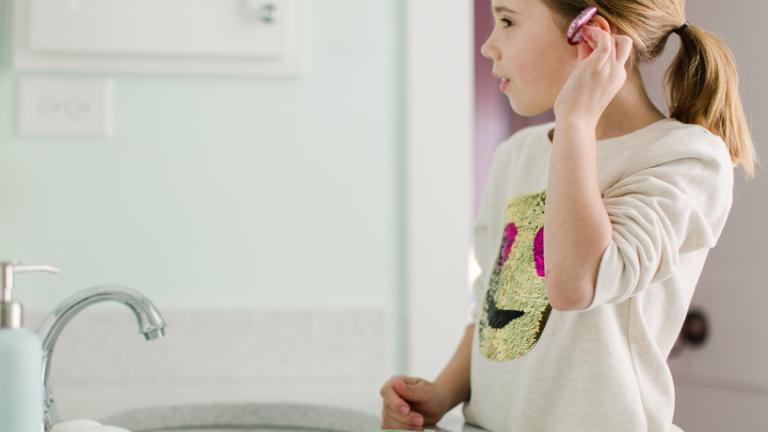 Image Description: a young girl fits her hearing aid into her ear while standing in front of her sink mirror.