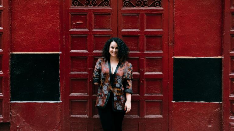 A young woman stands in front of a red background wearing a fashionable patterned jacket. One arm of the jacket is amended to fit her needs.