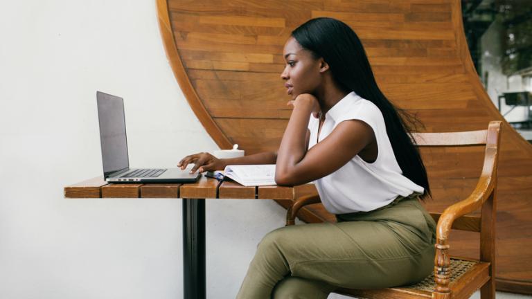 A woman sits at a table looking at her laptop.