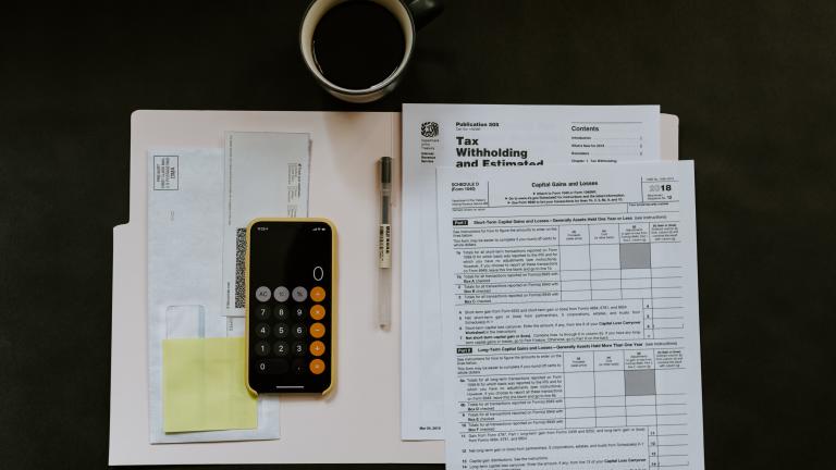 Image Description: Bird's eye view of tax forms, a calculator, and a cup of coffee displayed on the top a desk.