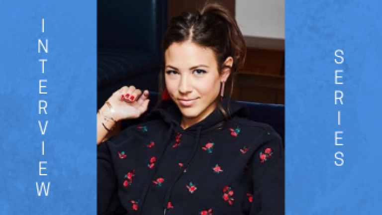 Image of a woman with brown hair in a ponytail smiling/smirking at the camera. She is wearing a navy button down shirt that has pink flowers on it.  She is wearing silver bracelets on her right arm, which is poised ever so slightly in an upward manner, while leaning on a navy chair.
