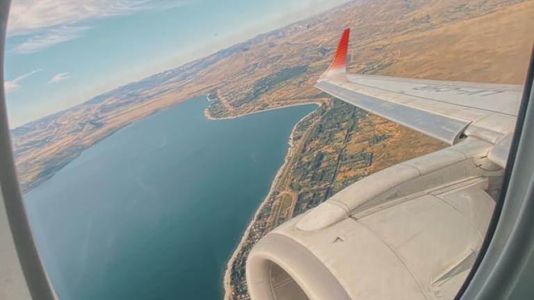Image of inside airplane window showing a birds eye view of undisclosed land and water below; it is angled and the airplane wing and engine is also featured. The sky in the photo is in daytime mode and there are several cirrus clouds floating.