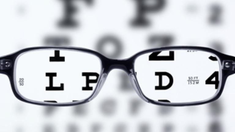 Image of an eye test chart which is blurry. There are black reading glasses superimposed on the image and through those lenses the letters L, D, P, and the number 4 can be seen.