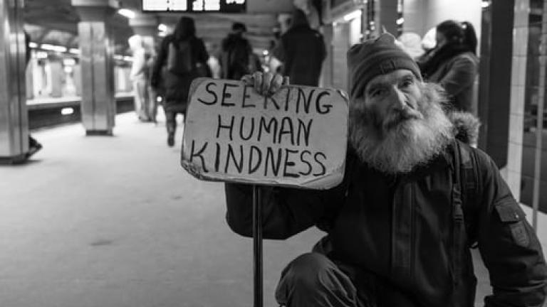 Homeless man in subway station holding a sign that says, &quot;Seeking Human Kindness.&quot; The photo is in black &amp; white.