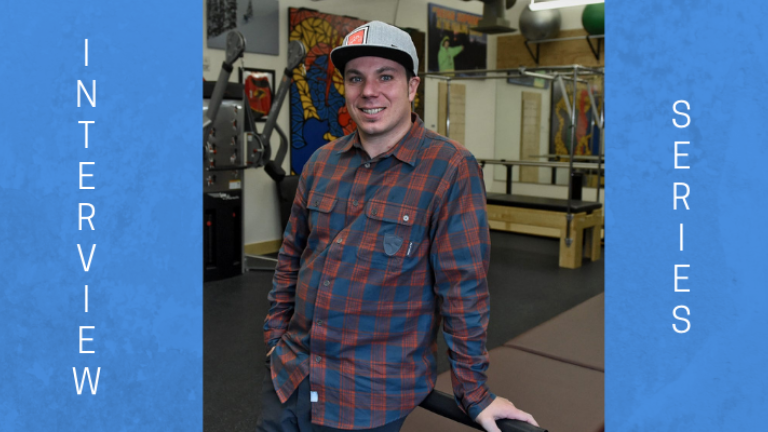 Image of Roy Tuscany. He is wearing a blue and red plaid shirt and jeans. His right hand is in his pocket. His left hand is on a table, which he is leaning against. He is Caucasian and is wearing a baseball hat and smiling at the camera. Roy has dark brown hair, which is protruding out of his hat. The background is of a physical therapy studio.